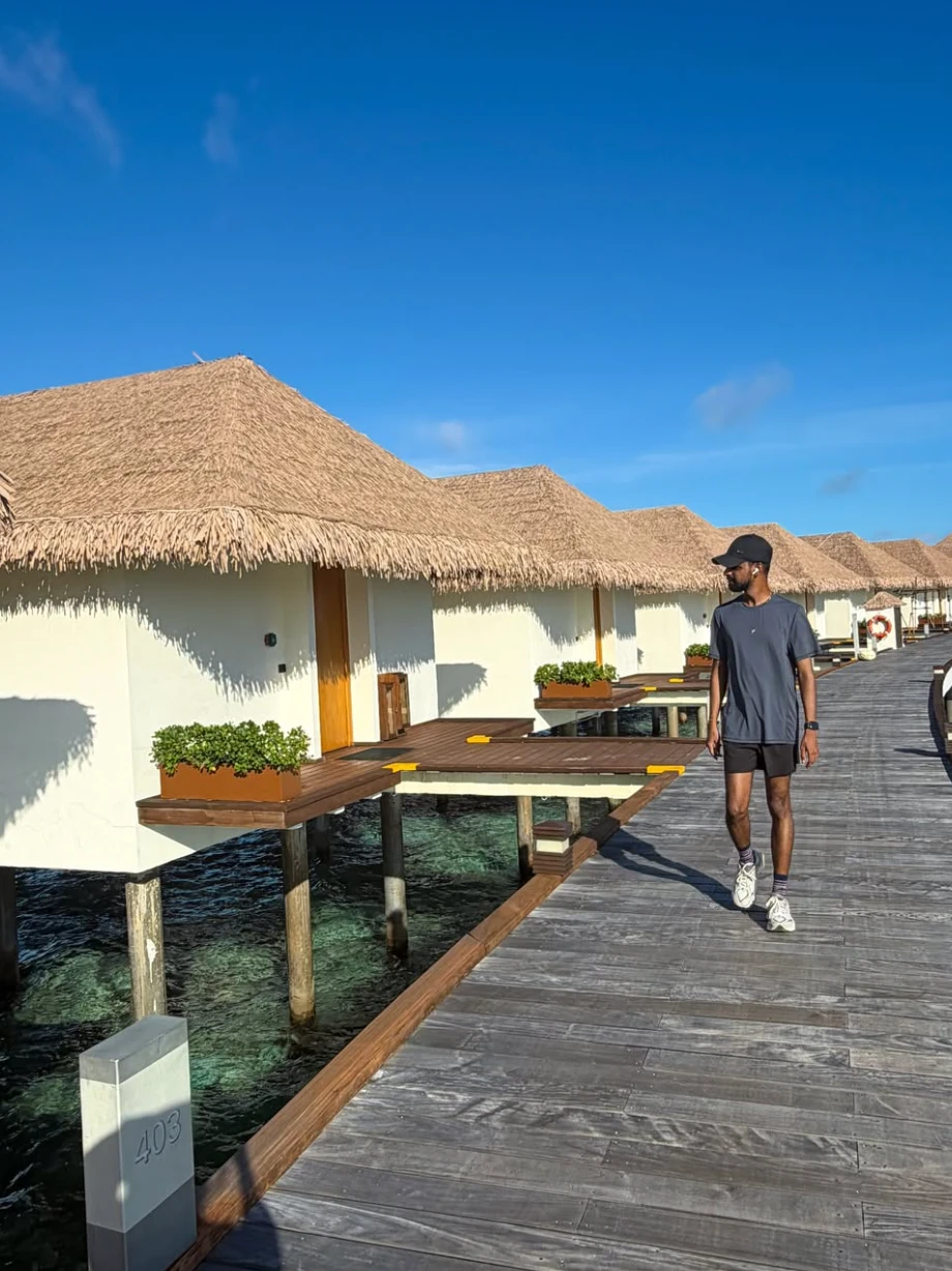 Dulanjaya walking along a wooden boardwalk past thatched over-water villas under a blue sky