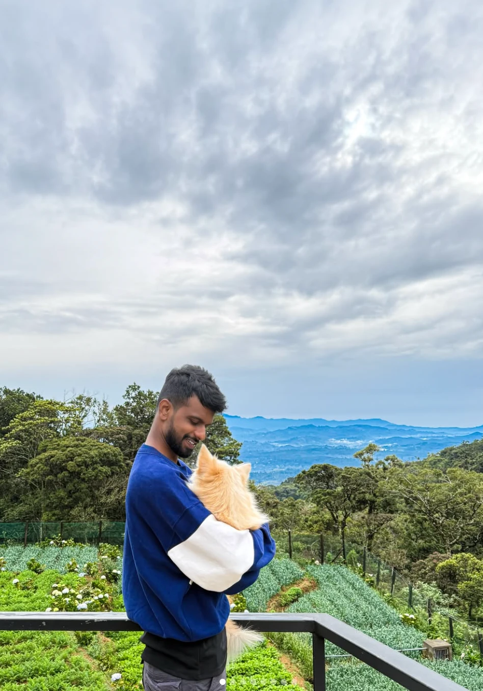 Dulanjaya smiling and holding a small cream-coloured fluffy dog on a viewpoint above green hills and misty mountains