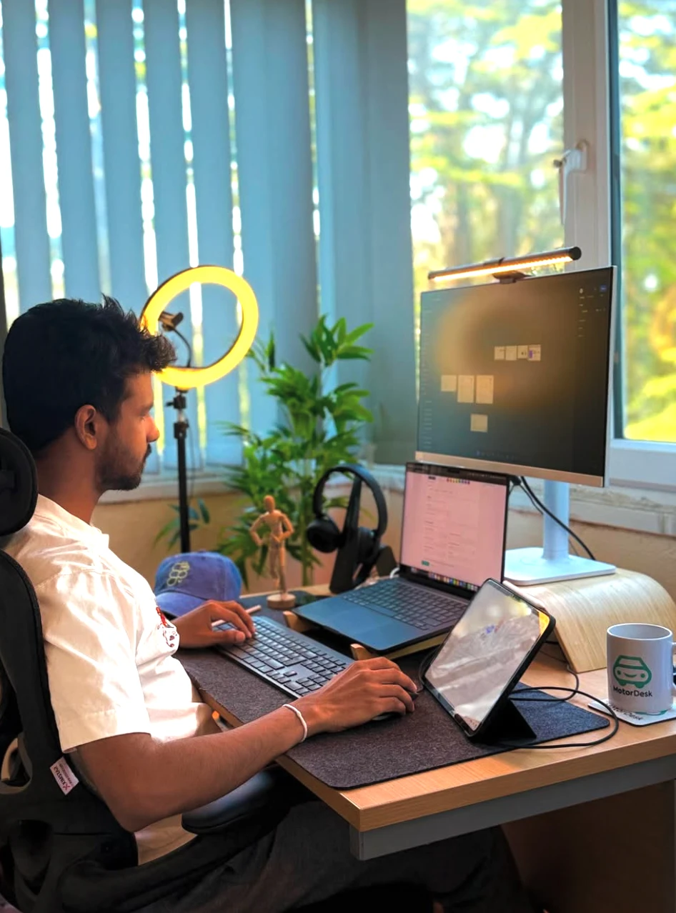 Dulanjaya working at a desk with a laptop, monitor, tablet and a MotorDesk mug by a sunny window